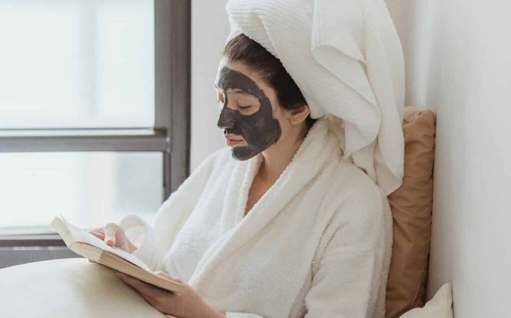 A women wearing a charcoal face mask sits in front of a window reading a book