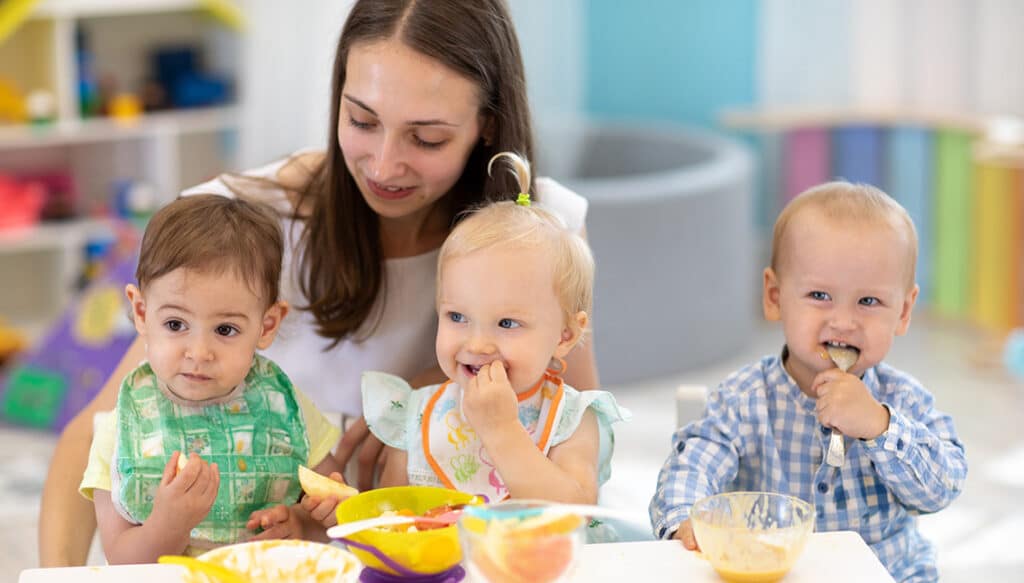 Infant Care qualified Nanny sitting at a table helping feed three smiling infants. Two boys and one girl.