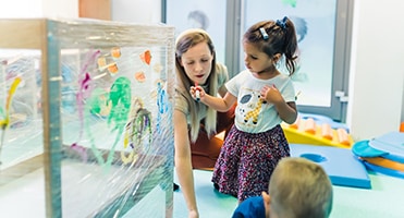 Cling film painting for improving kids imagination. Toddlers painting with, brushes, rollers and paints on a cling film wrapped around the wooden shelving stand