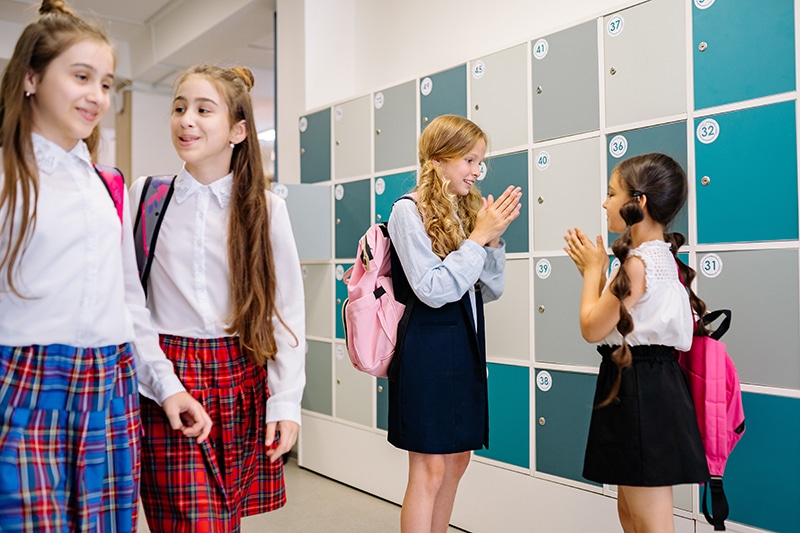 A group of girls walk by talking as two girls talk and play a game in a school hallway