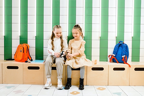 Two girls sit together on light-color bench outside of classroom playing together