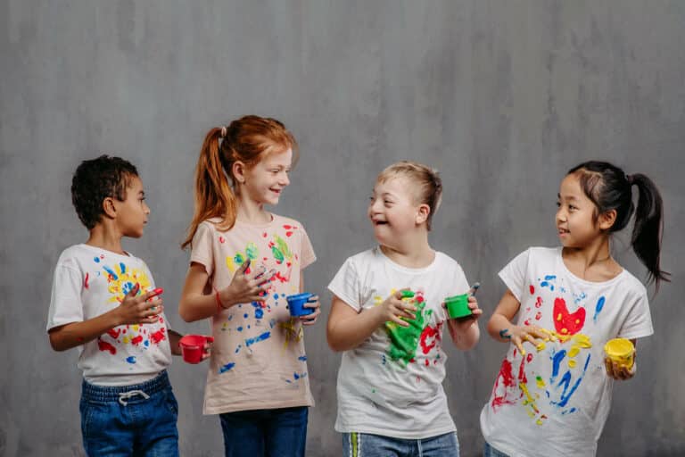 A group of children, one with down syndrome, stand in front of a gray backdrop smiling at each other with paint on their hands and shirts
