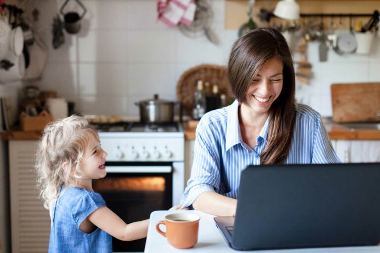 A happy mom managing work on her laptop while her daughter with a developmental delay laughs and pulls at her sleeve.